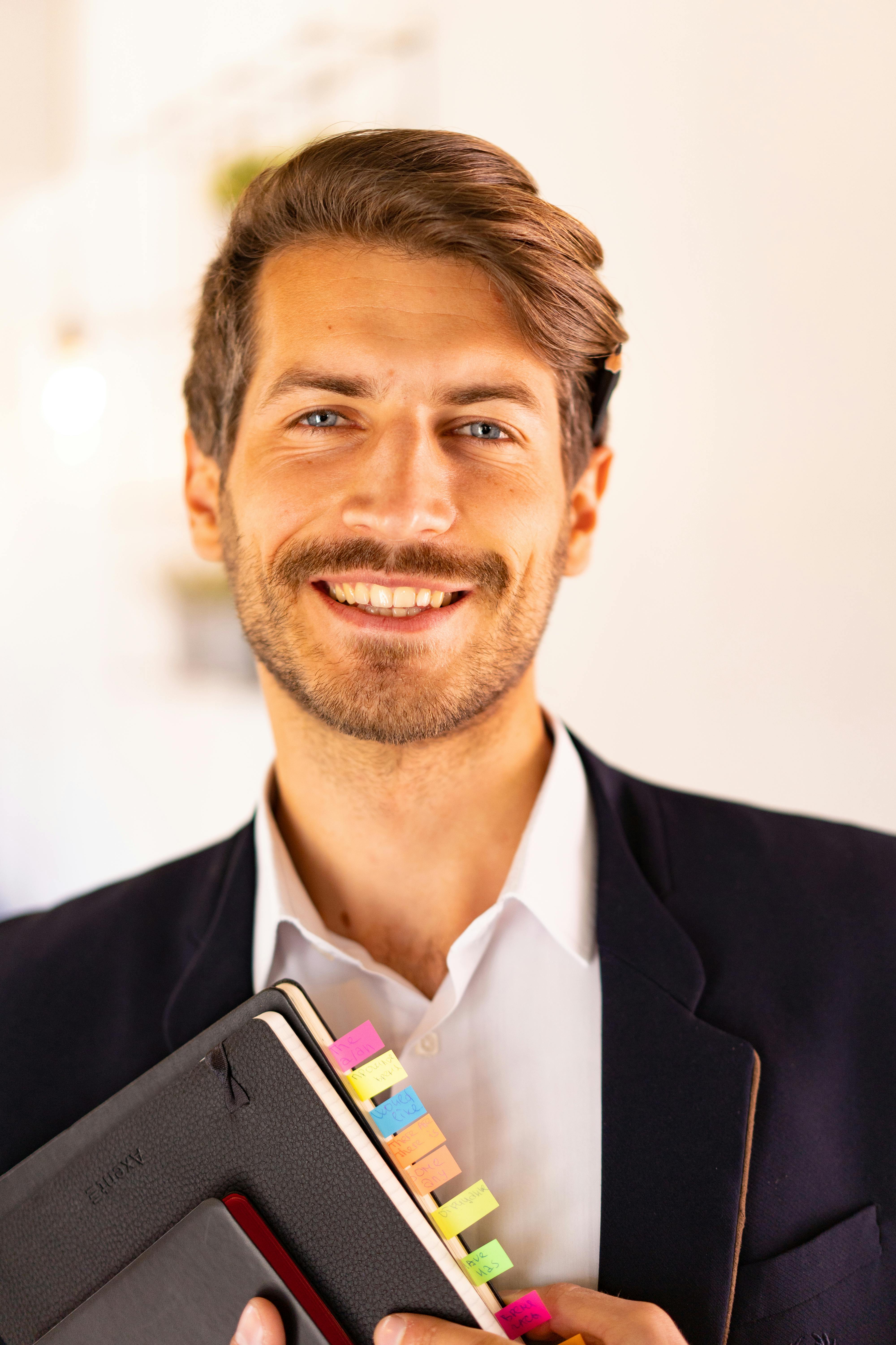 Smiling man in a suit holding colorful tabbed notebooks in a professional indoor setting.