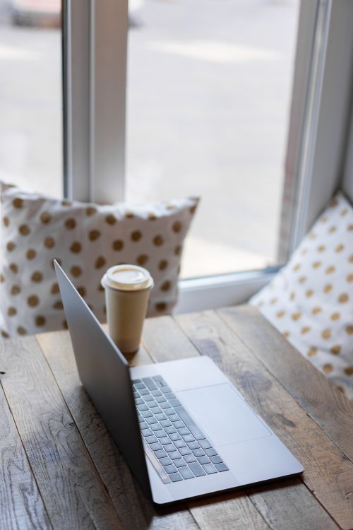 Free A serene workspace setup featuring a laptop and coffee beside a window with soft light filtering in. Stock Photo