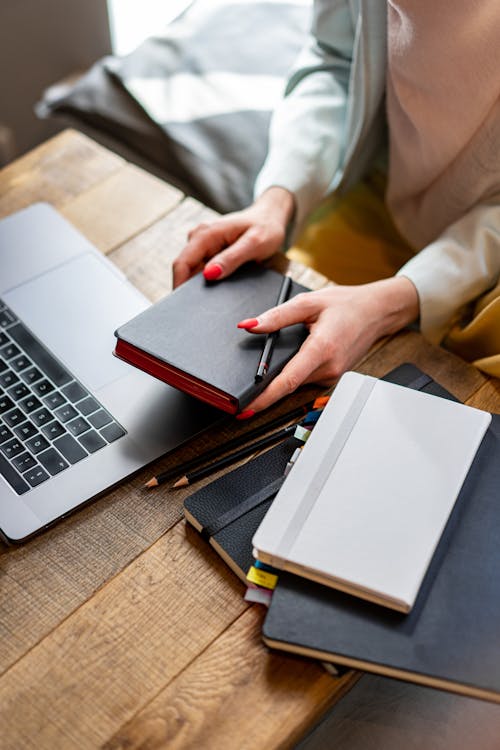 Woman's Hand Holding a Pen and Notebook Near a Laptop