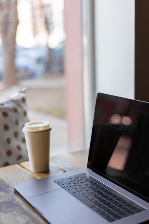 Free A cozy indoor scene featuring a laptop and disposable coffee cup on a wooden table by the window. Stock Photo