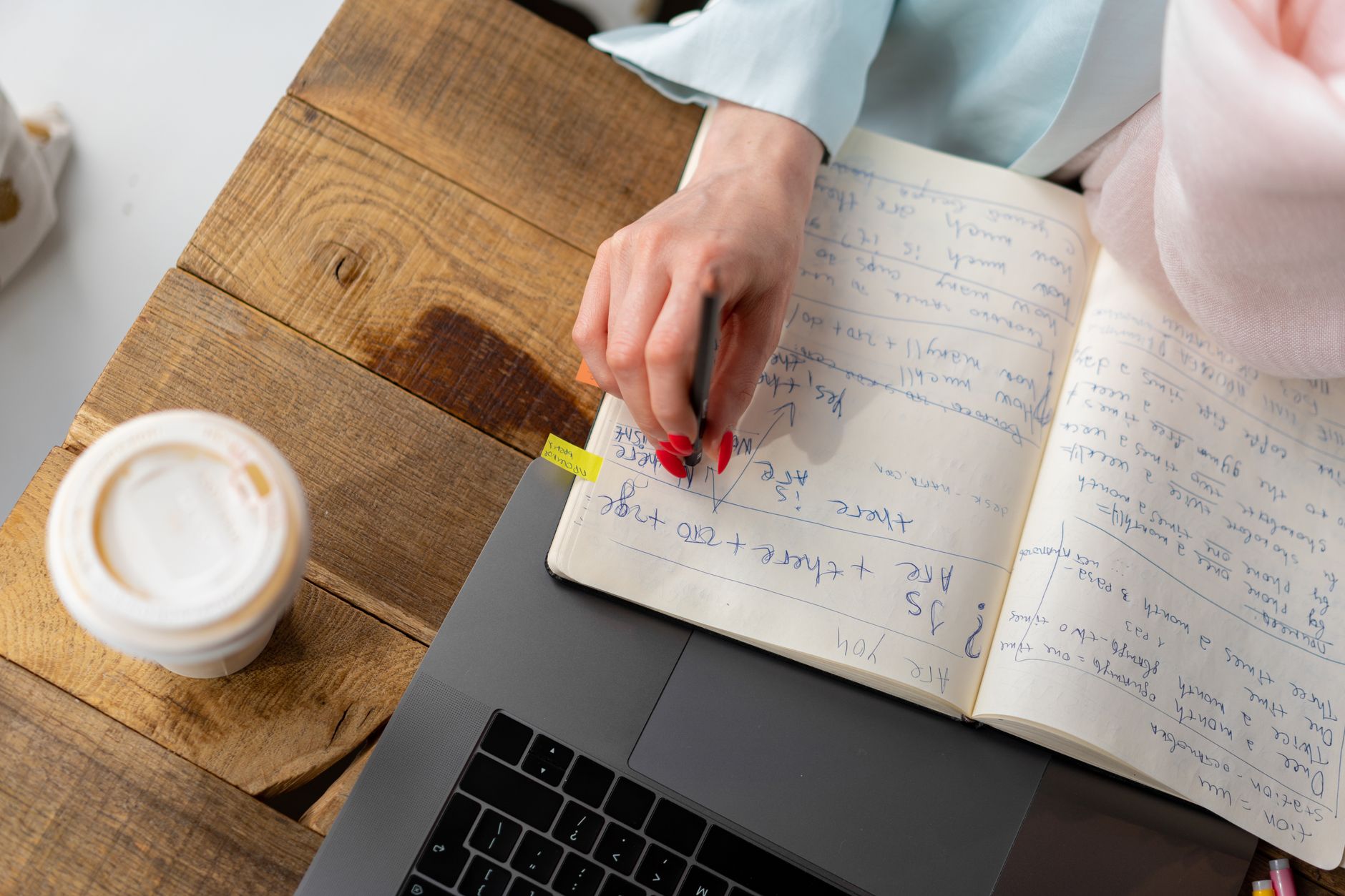 An open notebook of handwritten notes beside a laptop and a coffee cup on a wooden desk