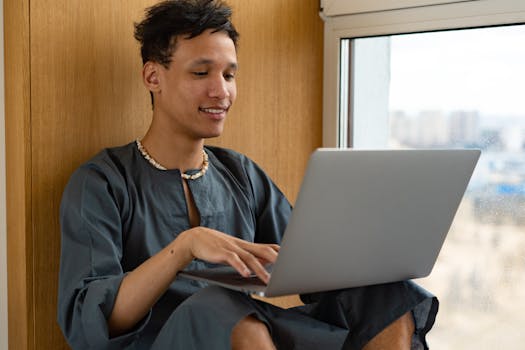 Young man in casual attire sitting by the window, using a laptop and smiling.