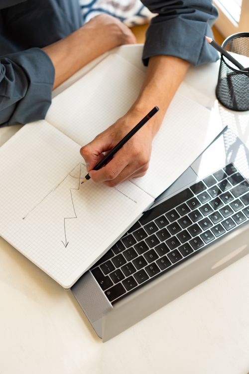 Free Person drawing a zigzag graph on a notebook placed atop a laptop; ideal for business or analysis themes. Stock Photo