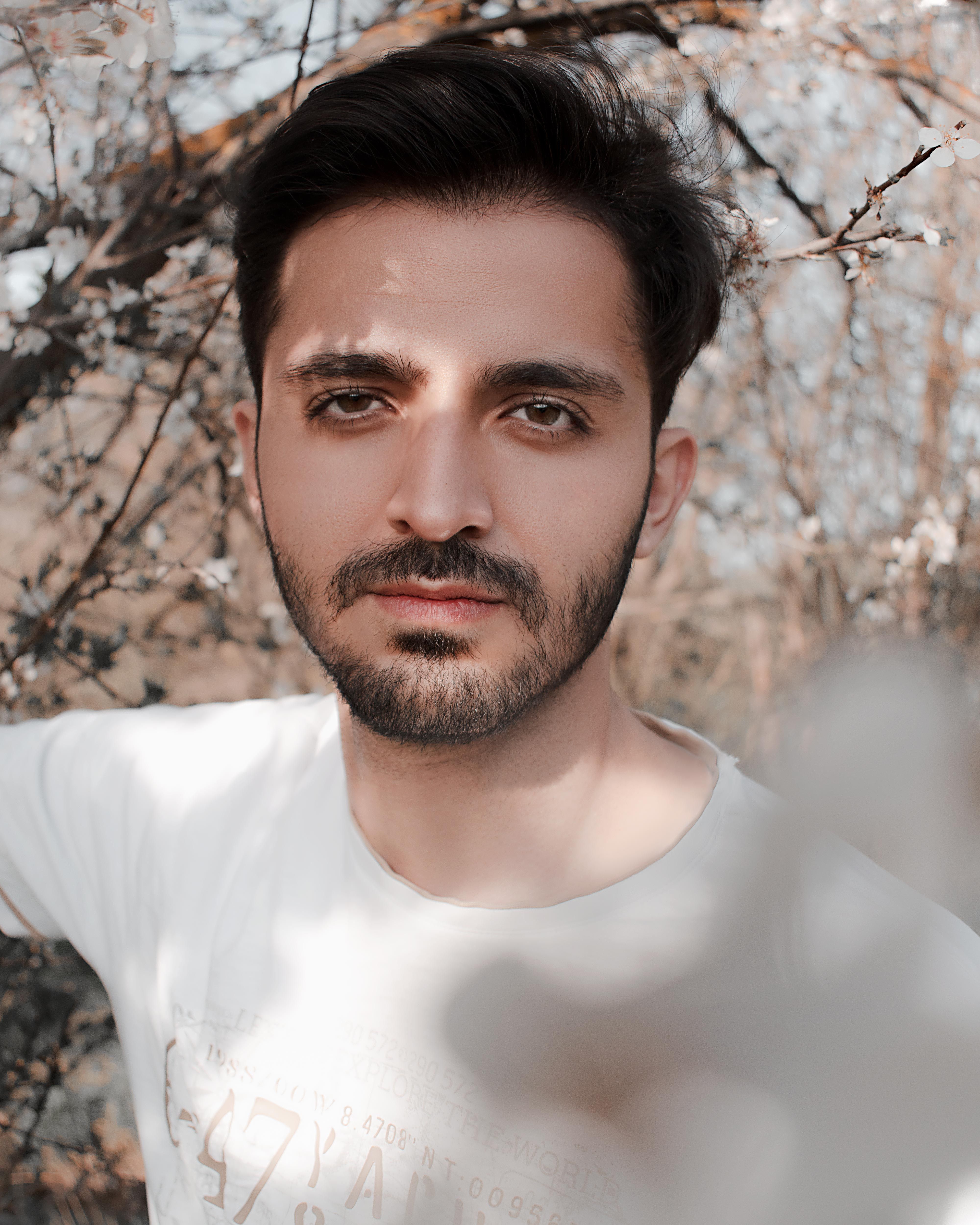 Close-up of a young man with facial hair standing outdoors among branches and blossoms.