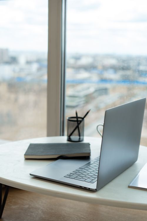 Free A sleek laptop and notebook on a bright workspace table by a window with city view. Stock Photo