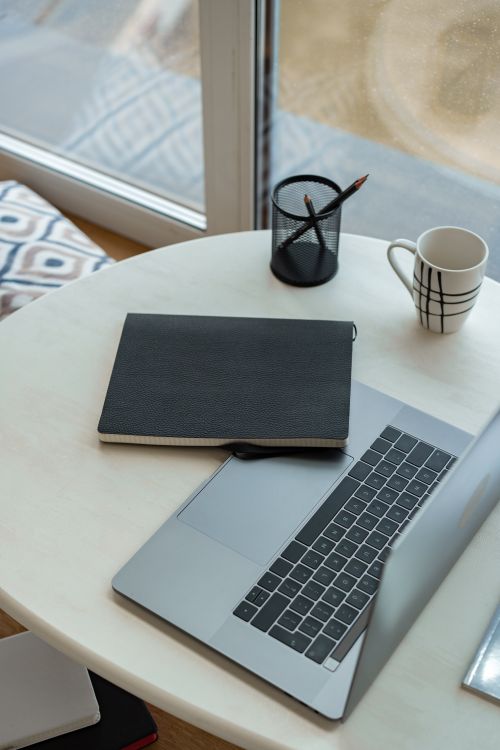 Free A stylish modern workspace featuring a laptop, notebook, pencil holder, and mug on a round table by a window. Stock Photo