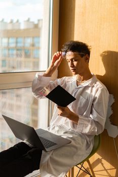 A young man in white attire reads by a bright window, laptop resting on his lap, with a thoughtful expression.