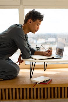 A young man works from home, writing notes beside a laptop with a cup of coffee on the table.