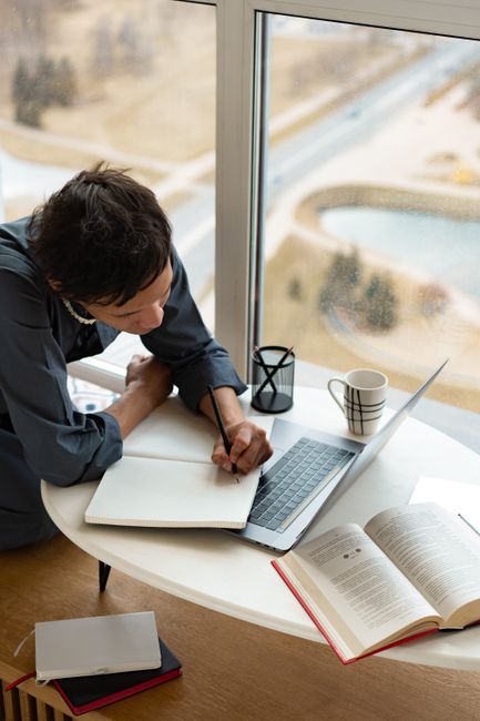 Adult man writing notes in notebook with laptop, book, and coffee cup on table by window.