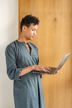 A young man in traditional attire using a laptop indoors, standing by a wooden door.