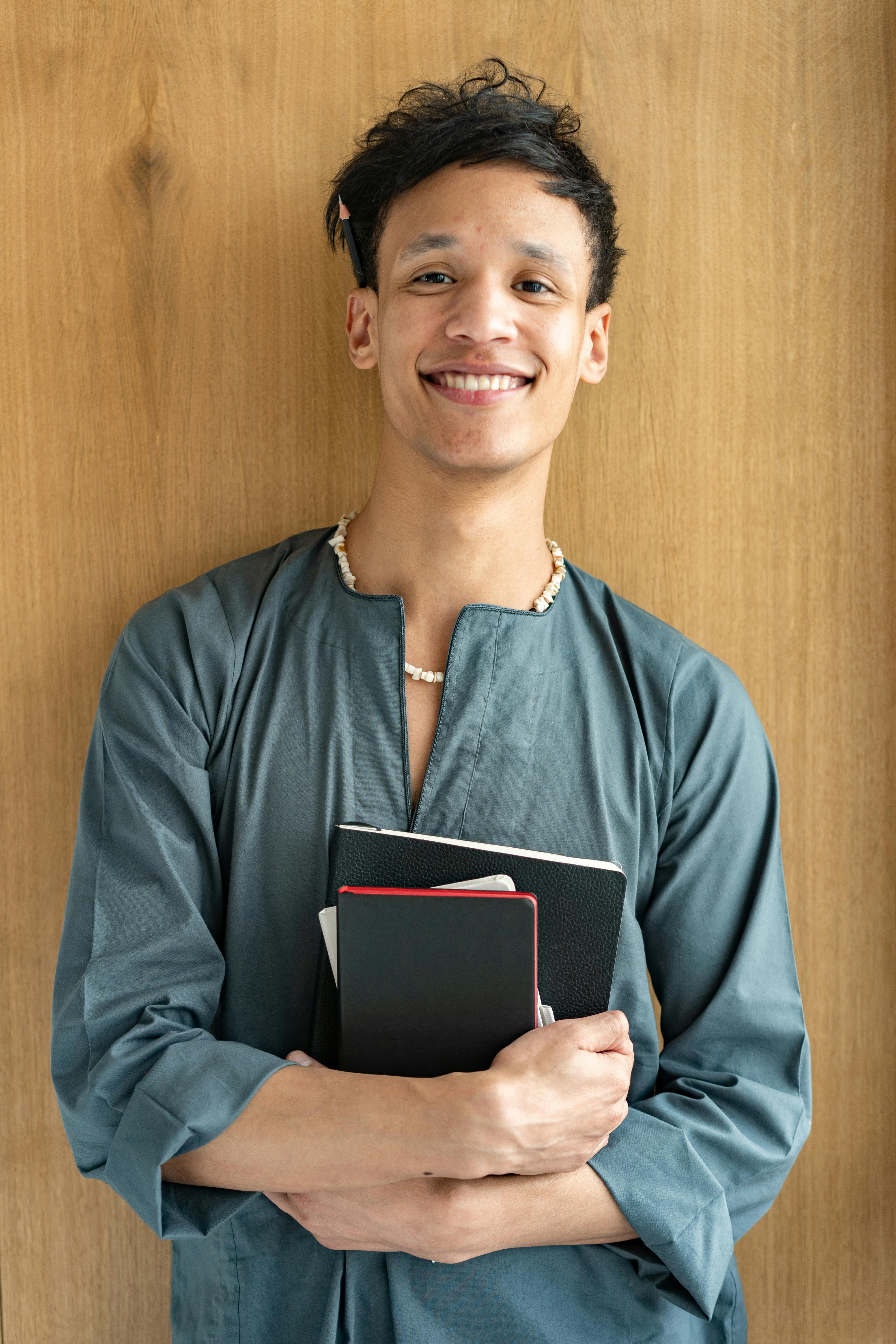 Young man smiling while holding notebooks, wearing casual attire against a wooden wall, exuding positivity.