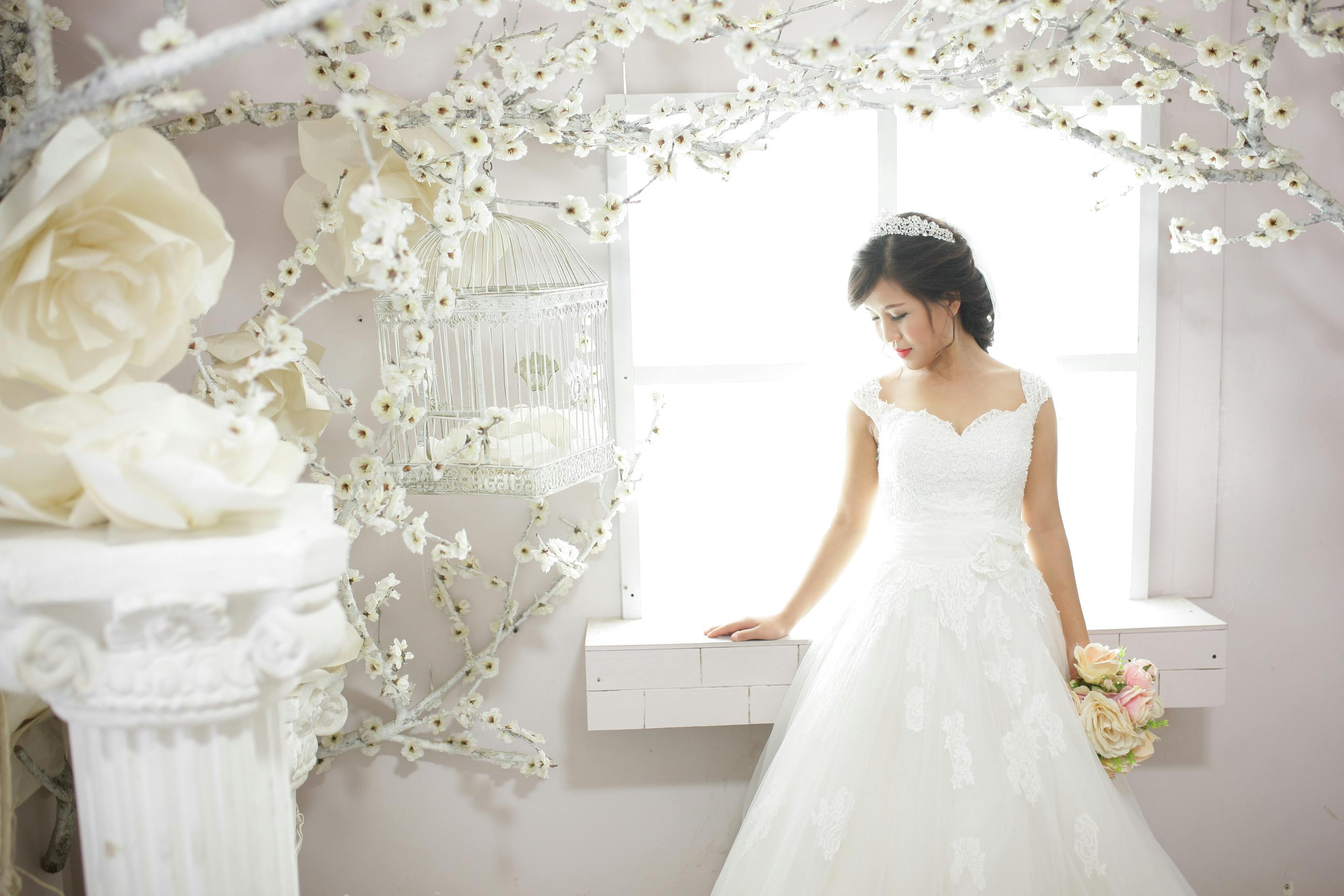 Beautiful bride in a white wedding dress holding a bouquet, standing by a bright window.