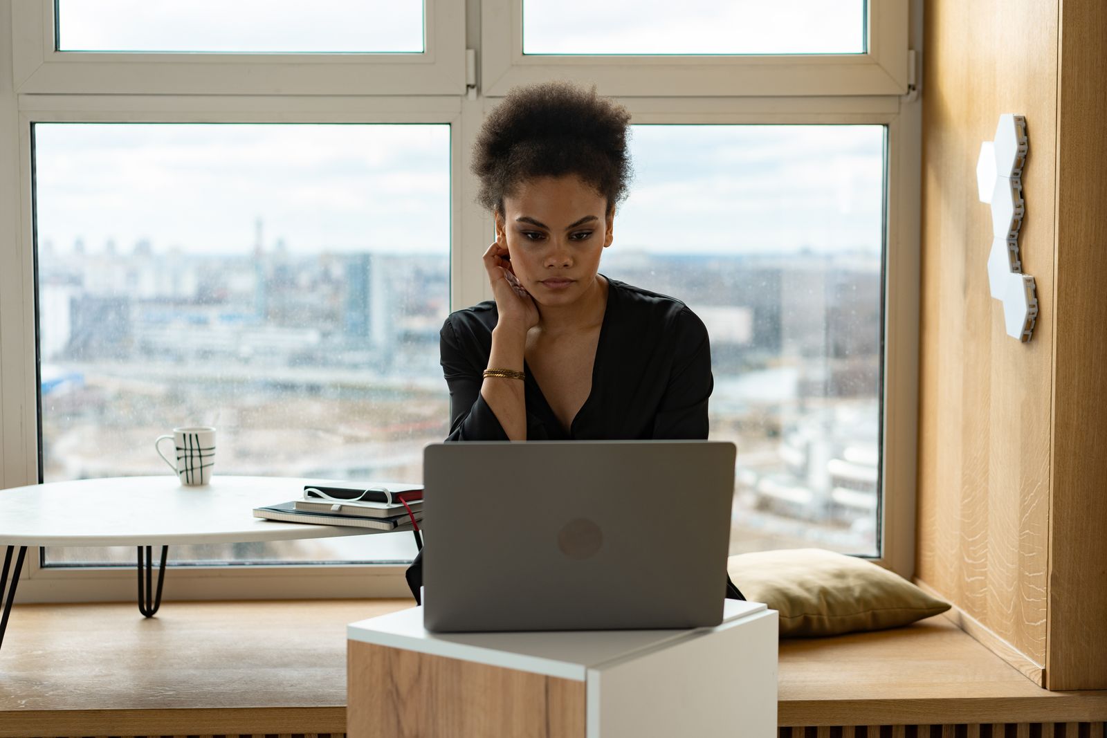 Woman in a black blouse working on a laptop beside a large window overlooking a city