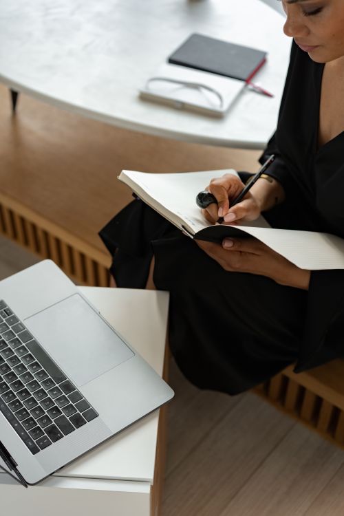 Free A woman writes notes in a notebook seated beside a laptop, capturing a modern work environment. Stock Photo