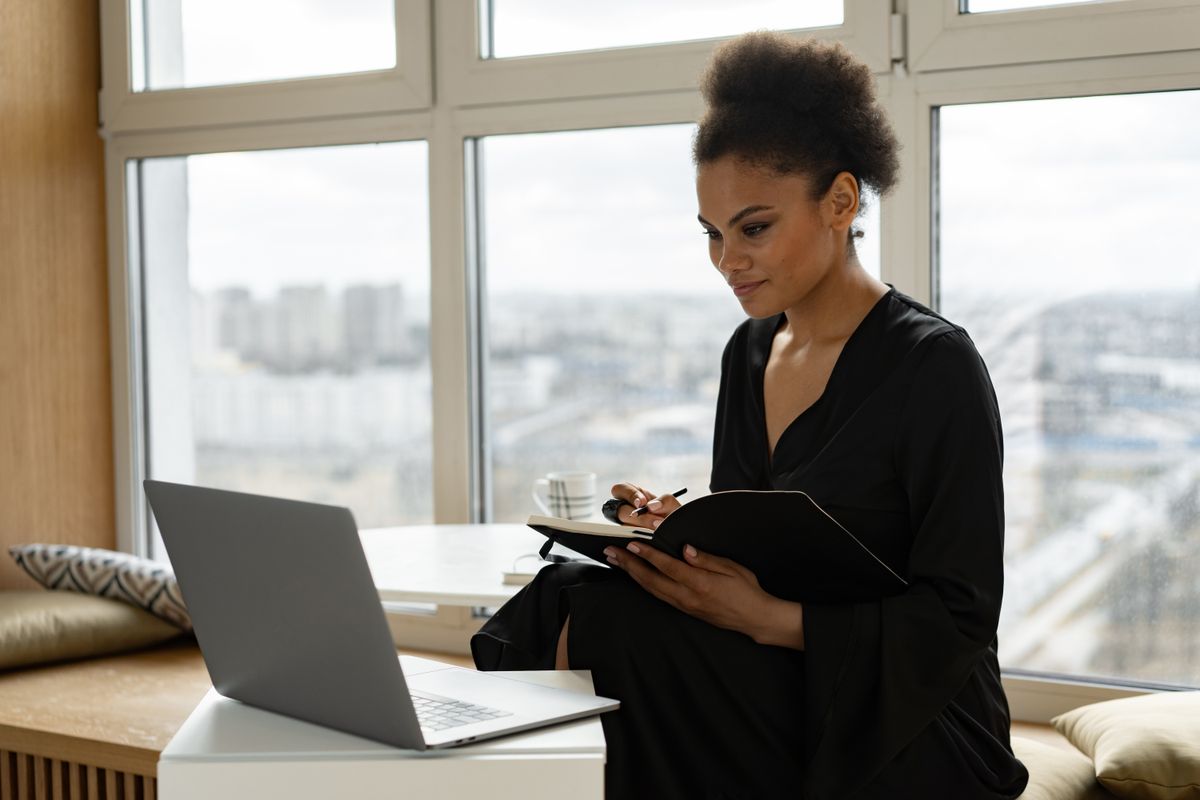 Black creative professional working with a laptop and notebook beside a window