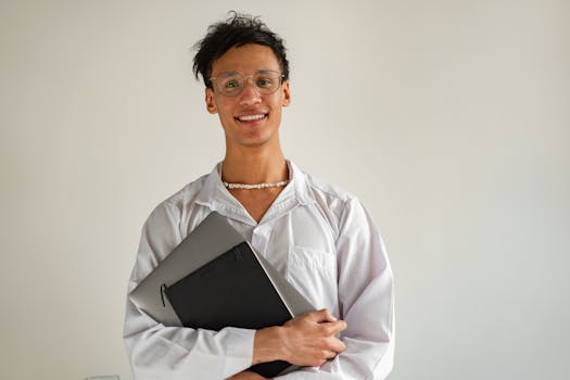 Young adult student with eyeglasses holding a laptop and notebook, standing indoors with a confident smile.