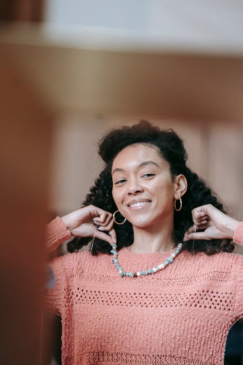 Cheerful African American female with black hair wearing pink sweater putting on mineral stone necklace while standing in light room at home