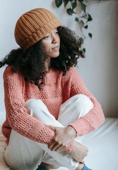 Young woman in a stylish knitted sweater and hat sitting calmly indoors, exuding warmth and tranquility.