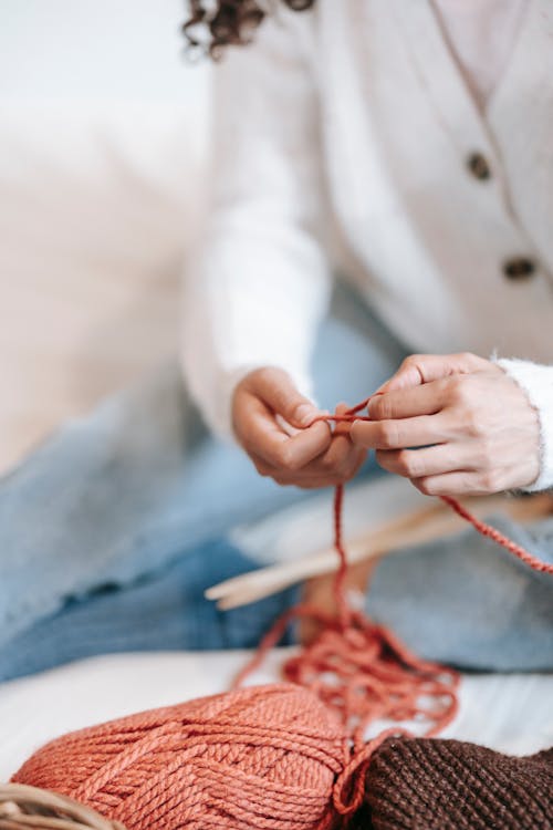 Crop anonymous female in casual wear with knitting needles and red threads sitting on bed in light room at home