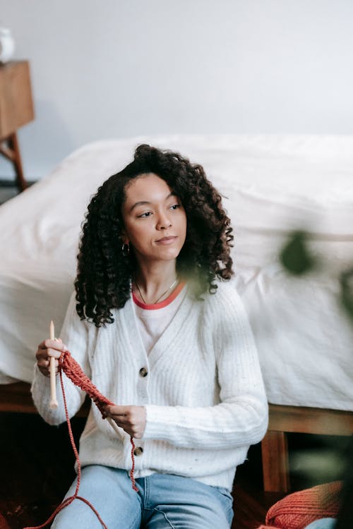 Talented African American craftswoman looking away while knitting with red yarn near white wall in light room with blurred green plants