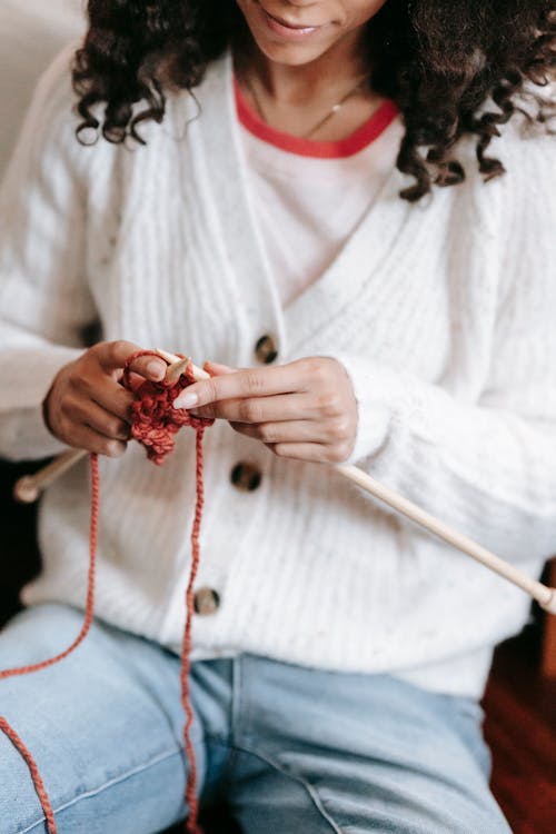 Crop faceless female in casual wear and black hair knitting with wooden needles while sitting in light room at home