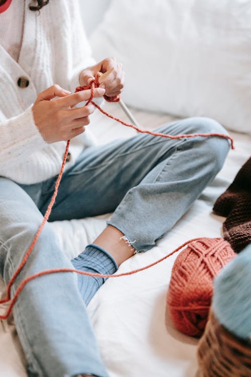 Crop unrecognizable talented female in jeans knitting with needles while sitting on white bed with skeins of threads in light room