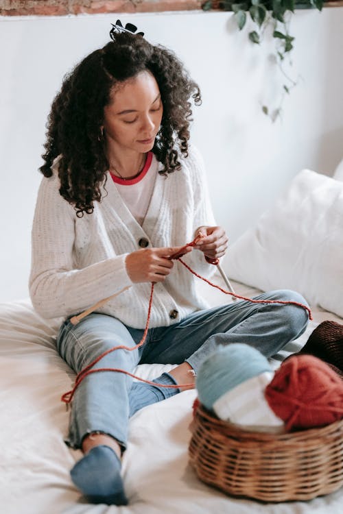 Full body of concentrated young ethnic female with curly in casual outfit sitting on bed and knitting with needles during weekend at home