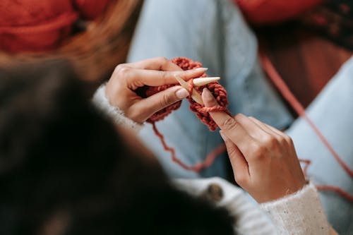 From above of unrecognizable female in warm clothes and jeans making stitches while knitting with yarn and needles sitting on floor at home