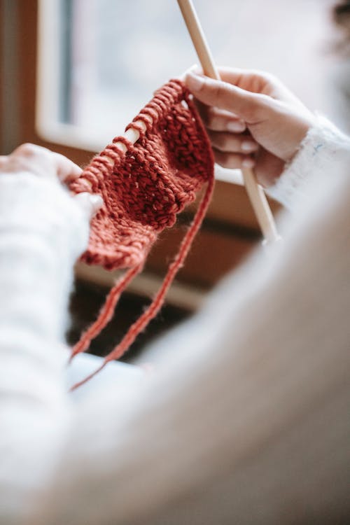High angle of crop anonymous lady in warm clothes knitting red scarf with wooden needles in daylight at home