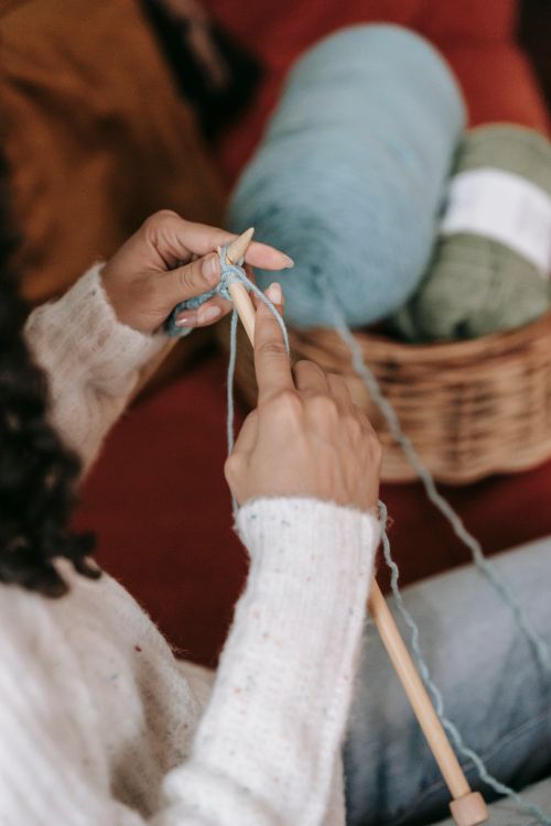 Free From above of crop faceless lady with dark hair in warm casual clothes sitting on cozy sofa with wicker basket with wool yarns and knitting with needles Stock Photo