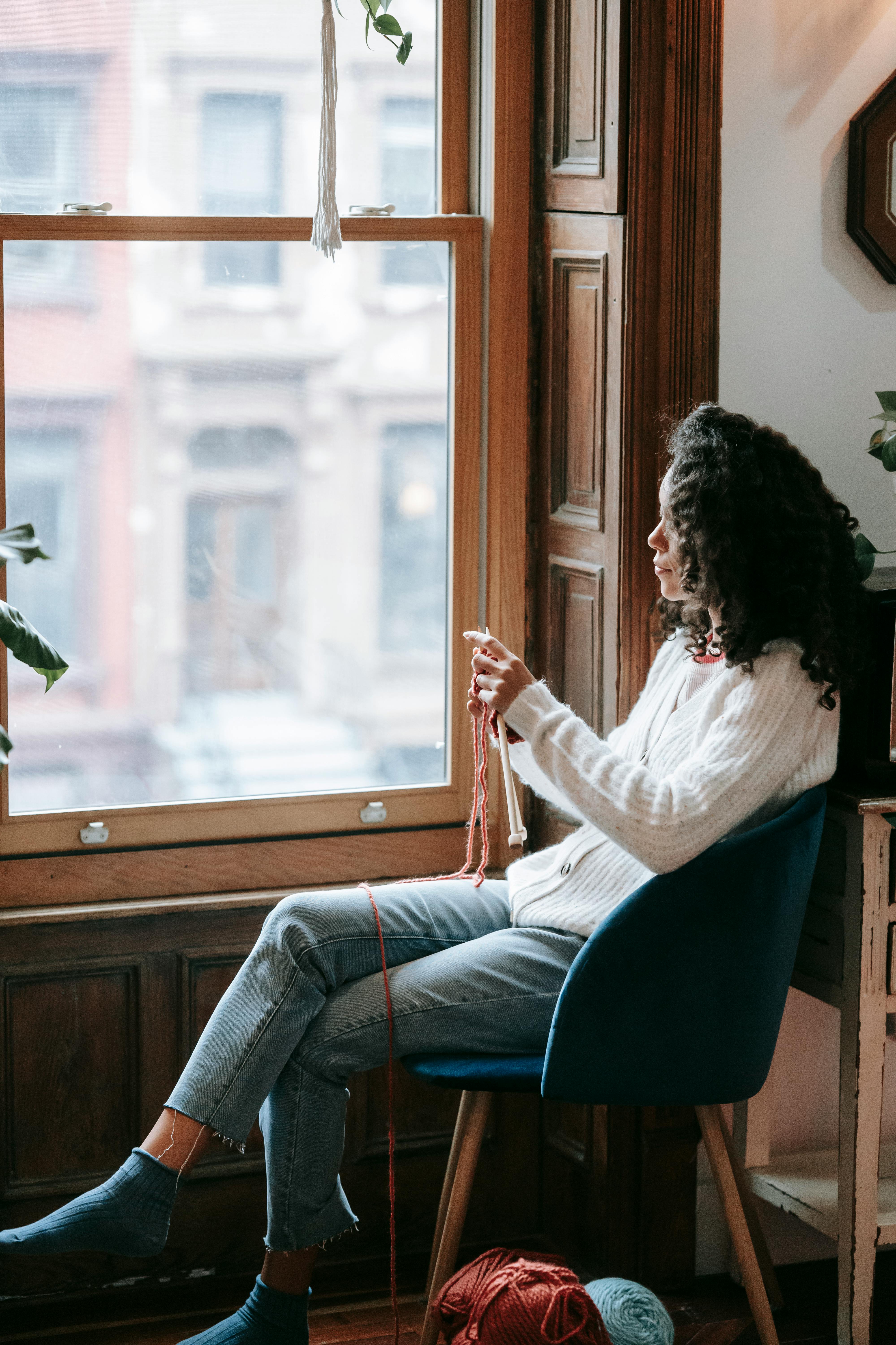 Side view of calm young ethnic female millennial with curly dark hair in casual outfit sitting on comfortable chair near window and knitting with needles in daylight