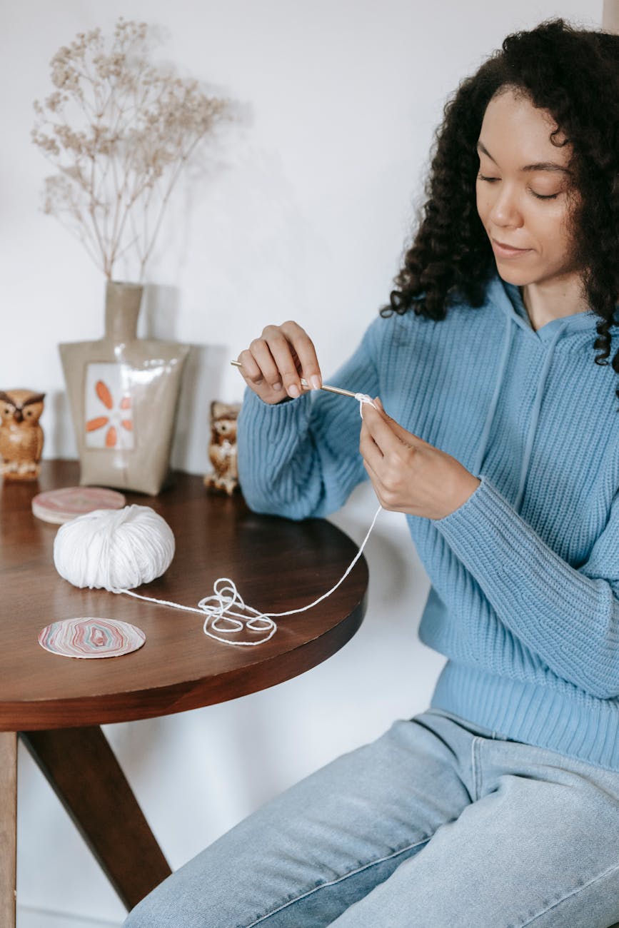 An adult woman crocheting at a wooden table, surrounded by yarn and cozy decor indoors.