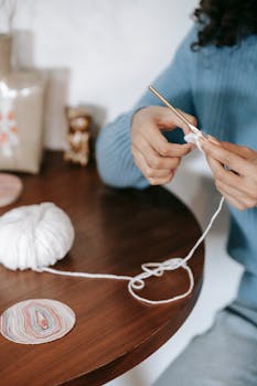 A woman in a blue sweater crochets with white yarn at a wooden table.