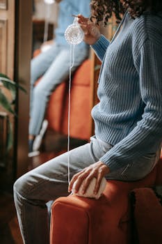 Adult crocheting with yarn and crochet hook on a sofa indoors, wearing a knitted sweater.