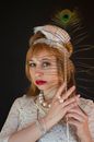 Attractive female model in white vintage dress with headband looking at camera with peacock feather in hand on black background