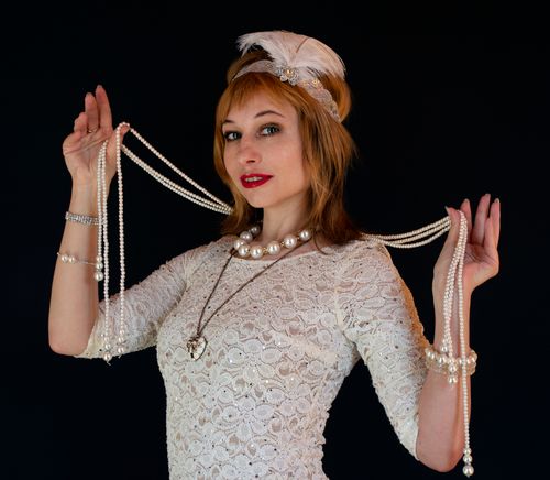 Free Charming female model with bijouterie in white vintage dress and headpiece with feather looking at camera on black background in studio Stock Photo