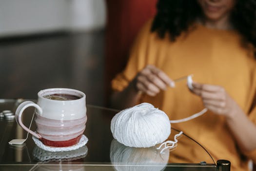 A cozy scene with tea and yarn as a woman crochets at a glass table.
