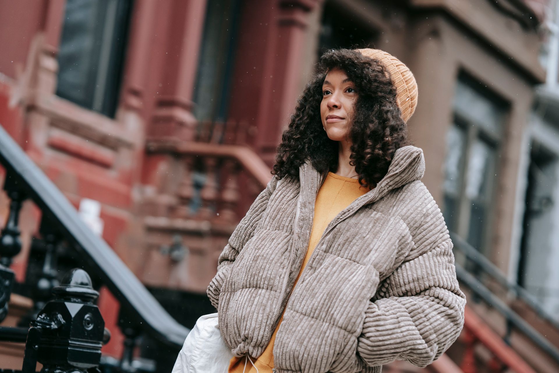 African American female with curly hair in outerwear with hands in pockets looking forward against urban building in cold weather