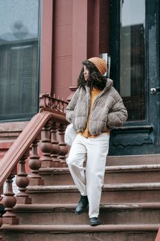 Trendy young African American female in outerwear with hands in pockets looking away while going down staircase in city