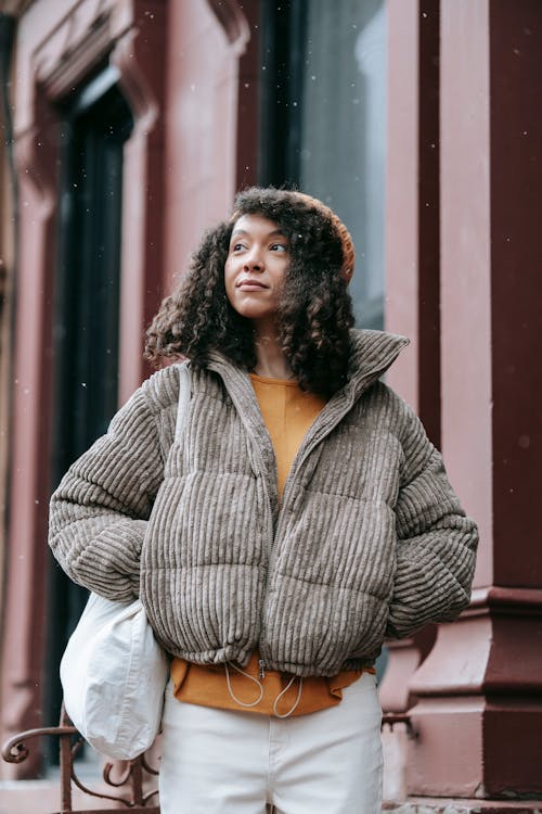 Contemplative young African American female in stylish outerwear with hands in pockets looking away in town