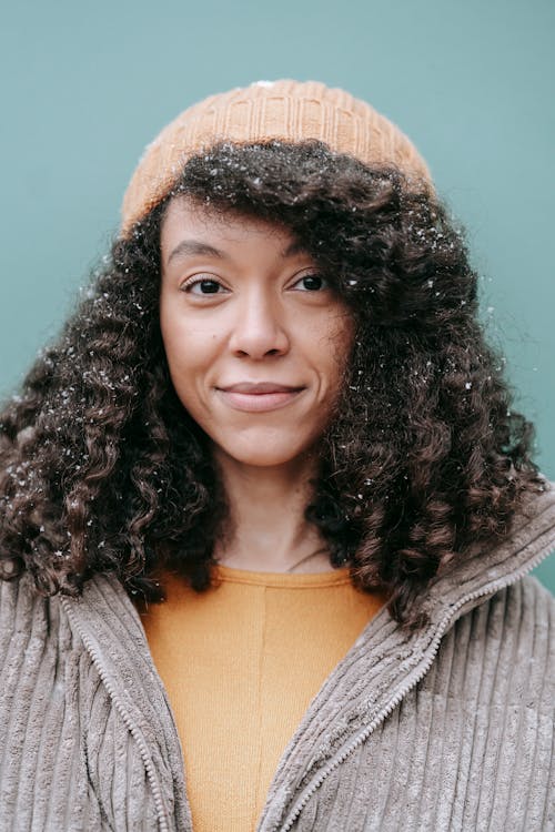Sincere young African American female in warm clothes with snowflakes on curly hair looking at camera on blue background