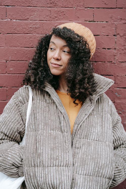 Dreamy young black female with curly hair in warm clothes standing with hands in pockets while looking away on street