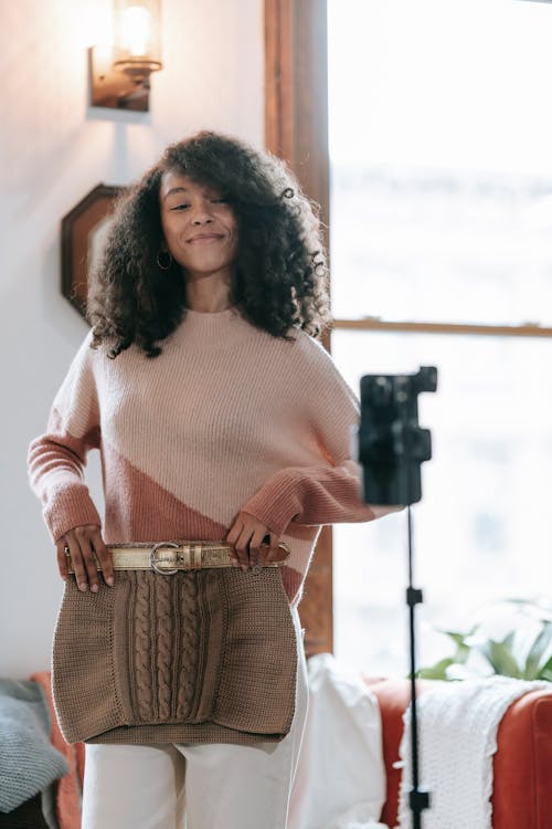 Cheerful young African American female vlogger with knitted skirt recording video on cellphone at home