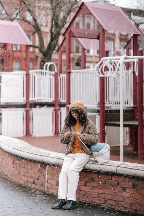 Full body of focused African American woman with curly hair in stylish clothes sitting on stone boarder on city street and knitting in winter time in daylight