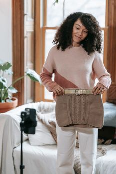 Cheerful African American woman with curly hair standing and demonstrating knitted skirt while recording video on smartphone in light room in daytime