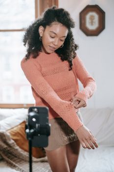Ethnic female blogger with curly hair showing knitted sweater to smartphone while filming advertisement for blog