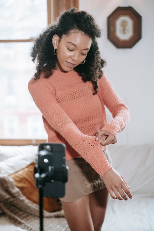 Free Ethnic female blogger with curly hair showing knitted sweater to smartphone while filming advertisement for blog Stock Photo