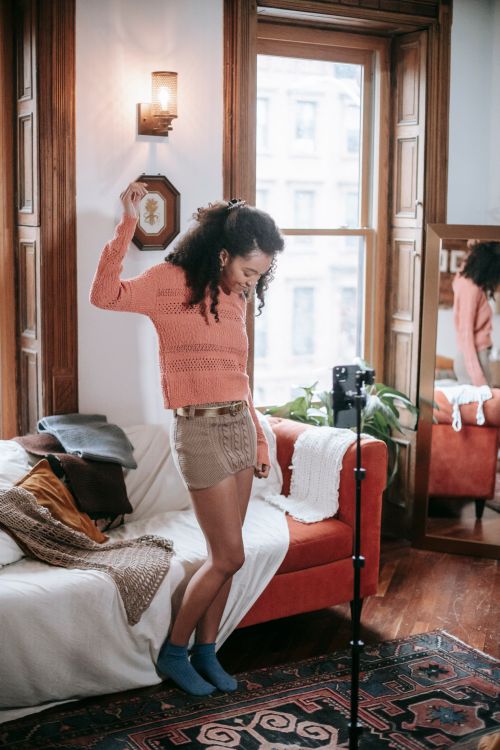 Free Full body of pleasant ethnic female blogger wearing knitted skirt and sweater standing with raised hand and filming video on cellphone on tripod Stock Photo