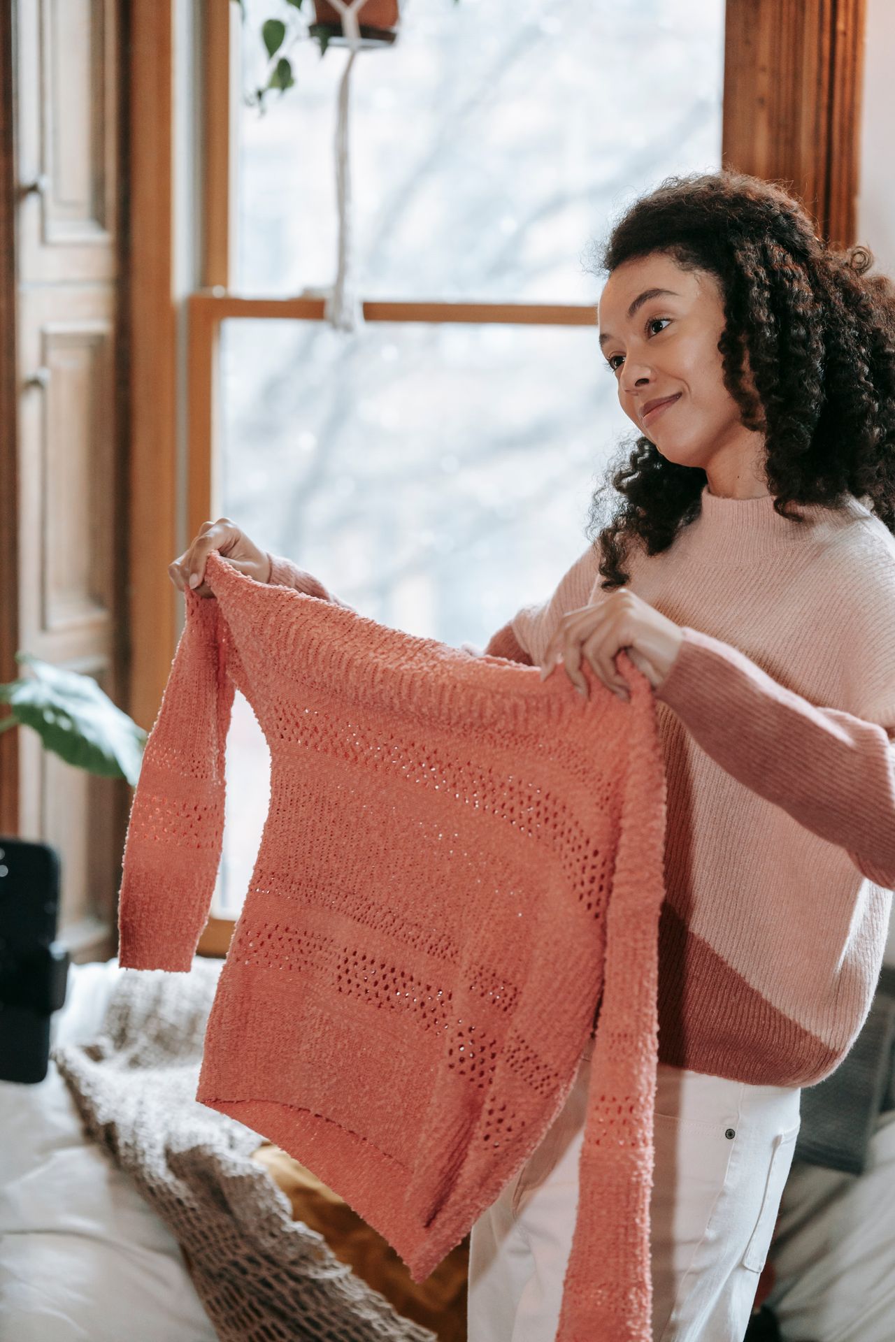 Woman in a pink sweater holding up a coral knit sweater by a window