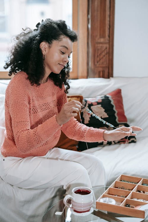 Free Side view of concentrated African American female with black hair stringing beads while making handmade bracelet while sitting in couch Stock Photo